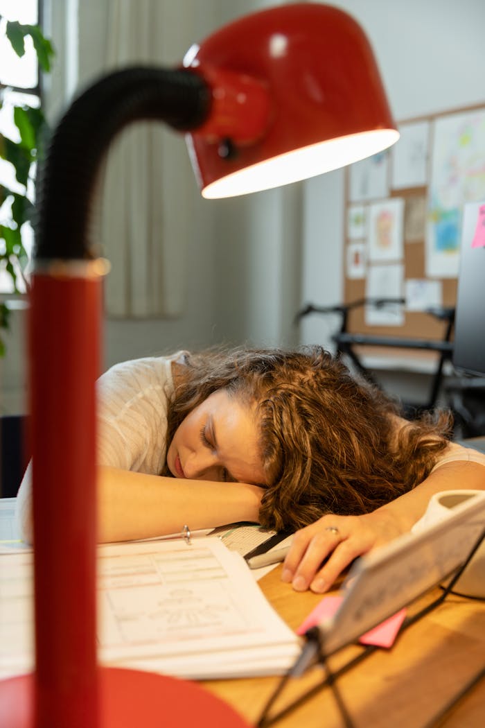 A businesswoman falls asleep at her desk, overwhelmed by work, under a red lamp.