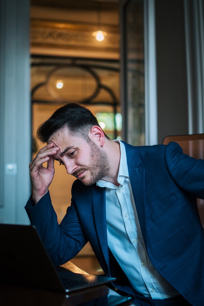 A worried businessman in a blue suit working on his laptop in an office environment.